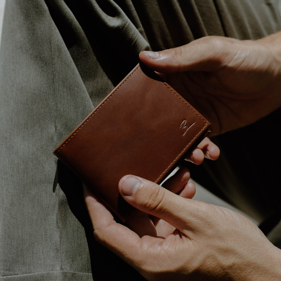 Three quarter view of Bullstrap Reserve Artisan bifold wallet in cognac leather with embossed B logo held against olive fabric