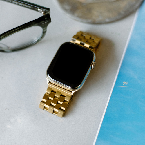Gold smartwatch with black screen on a light surface next to eyeglasses and a blue book showing the number 10