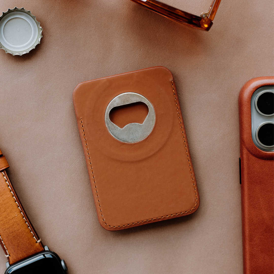 Brown leather MagSafe wallet with built-in metal bottle opener beside a leather watch strap and phone case