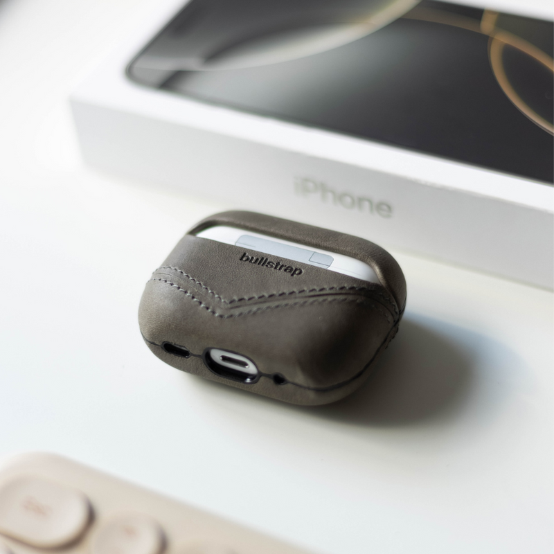 Leather AirPods case stamped bullstrap on white surface, iPhone box blurred in background