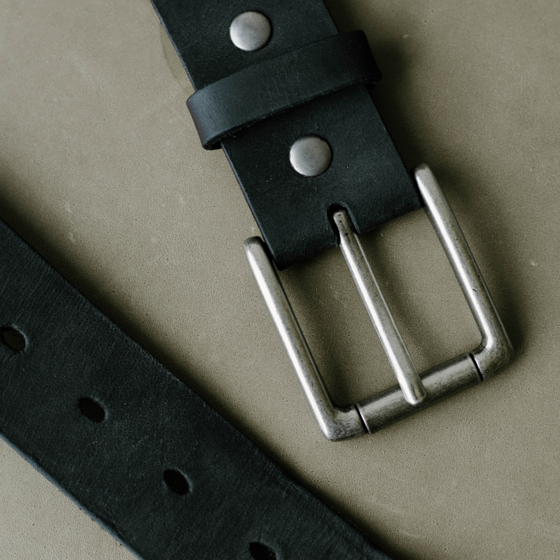 Close-up of Rugged Leather Belt Black Edition showing matte silver buckle, riveted black strap, and keeper on tan background