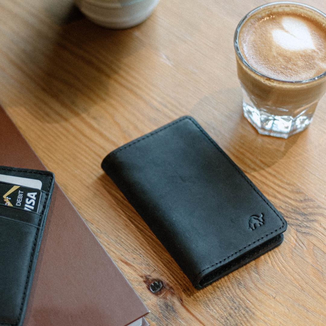 Black leather messenger wallet on wooden table beside coffee and cards showing DEBIT VISA
