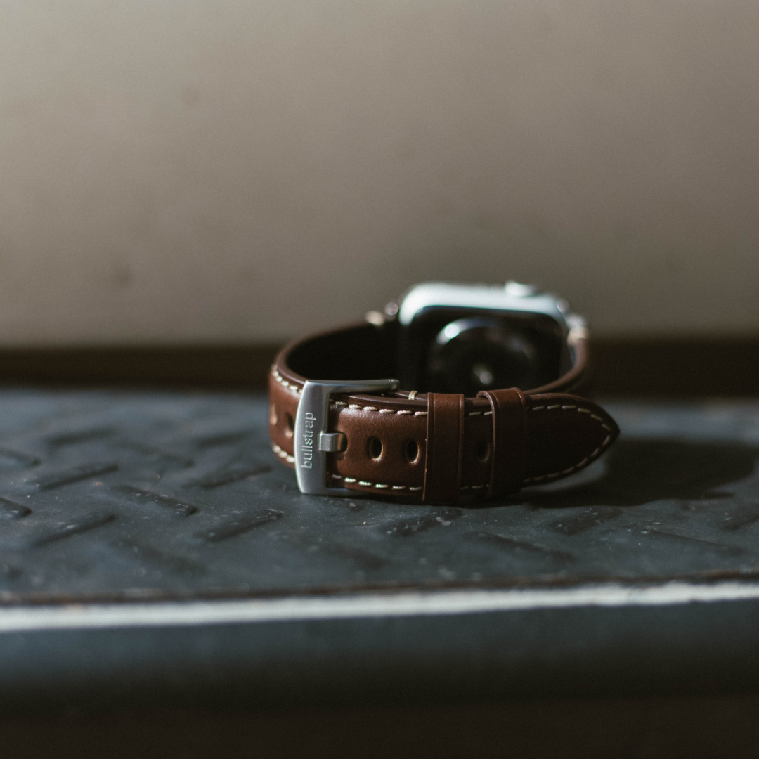 Brown leather watch strap with stitched edges and metal buckle engraved 'bullstrap,' watch body blurred in background.