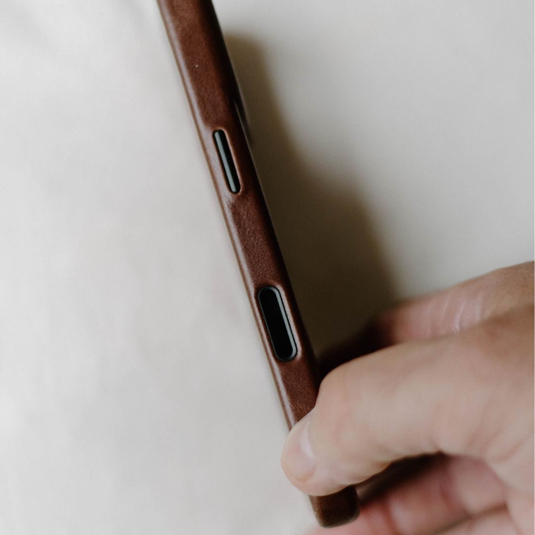 Brown leather phone case edge with button and port cutouts, held between thumb and fingers.