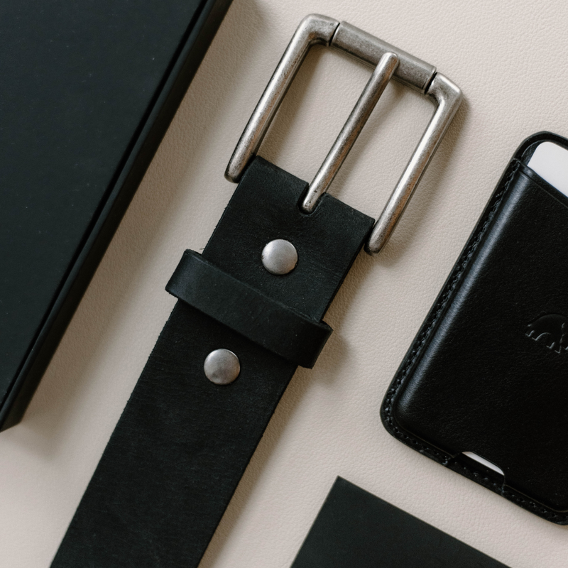 Close-up of Rugged Leather Belt Black Edition showing silver buckle and double-rivet strap against cream background