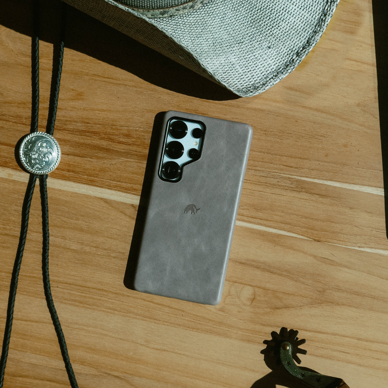 Smartphone with gray leather case on wooden table beside a straw cowboy hat, bolo tie concho, and a spur