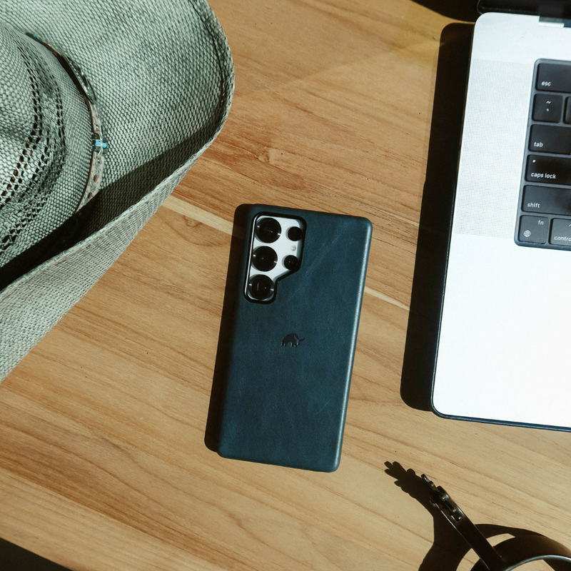 Straw hat, dark leather phone with multi-lens camera on wooden table beside laptop showing keys: esc, tab, caps lock, shift, fn, control.