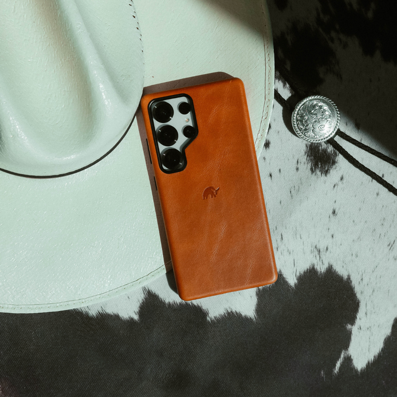 Smartphone with tan leather case resting on a white cowboy hat and black-and-white cowhide, silver bolo tie nearby.