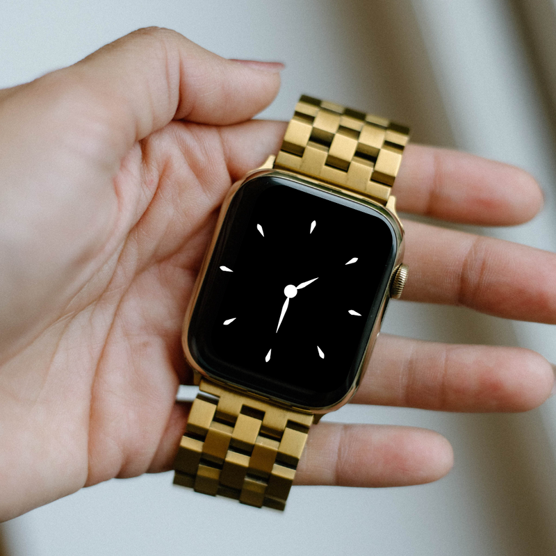 Gold metal Apple Watch with linked strap held in a hand, showing a black watch face with white clock hands and markers