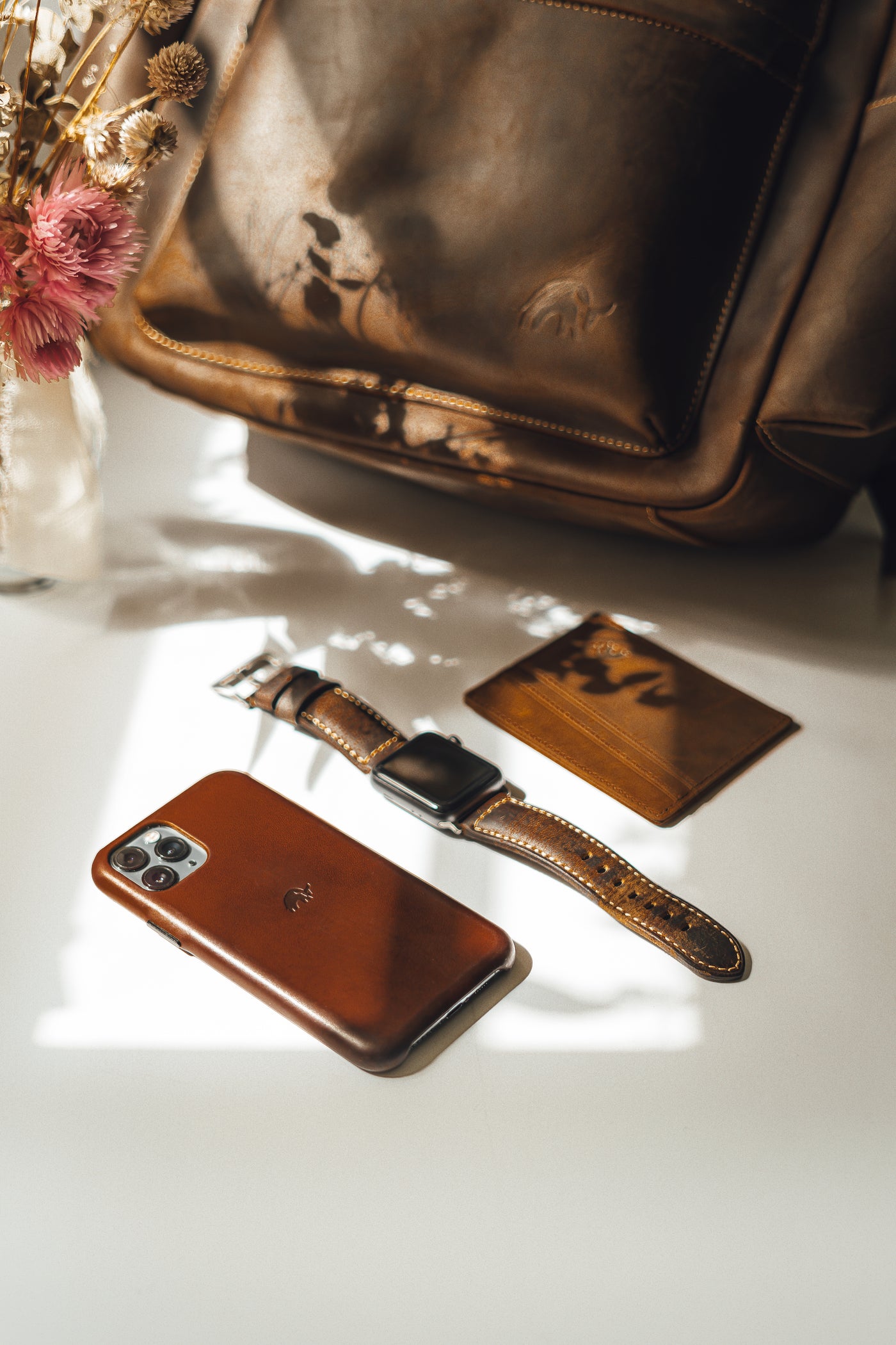 A leather phone case, watch, and cardholder on a table with a bag in the background.