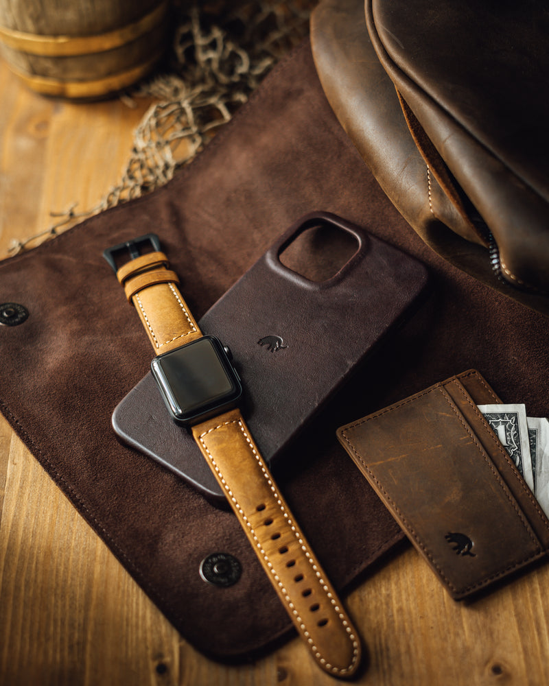 Brown leather phone case beneath a tan smartwatch band and slim wallet, with a dollar bill showing ONE on a wooden table.
