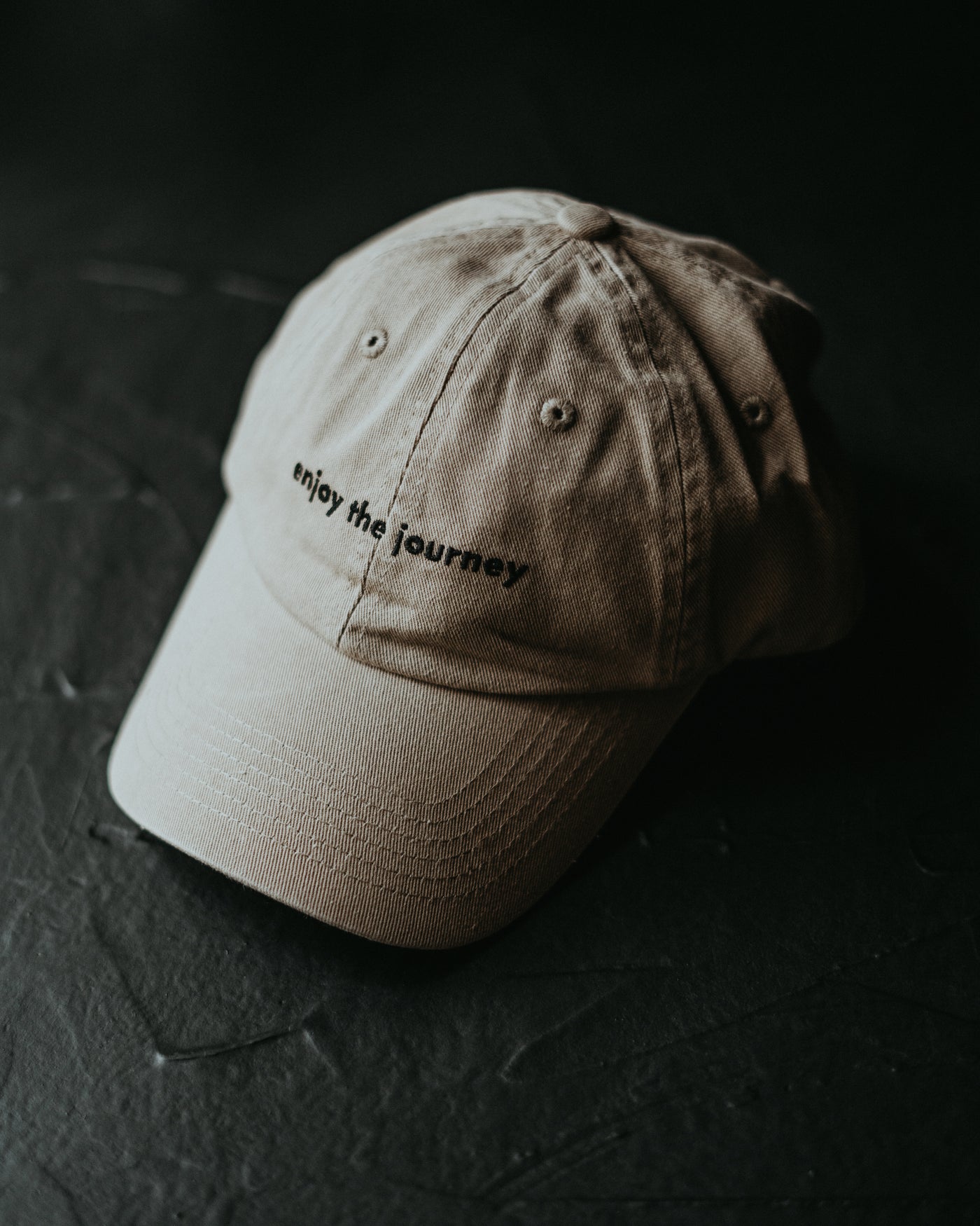 Beige baseball cap on dark surface, embroidered with enjoy the journey