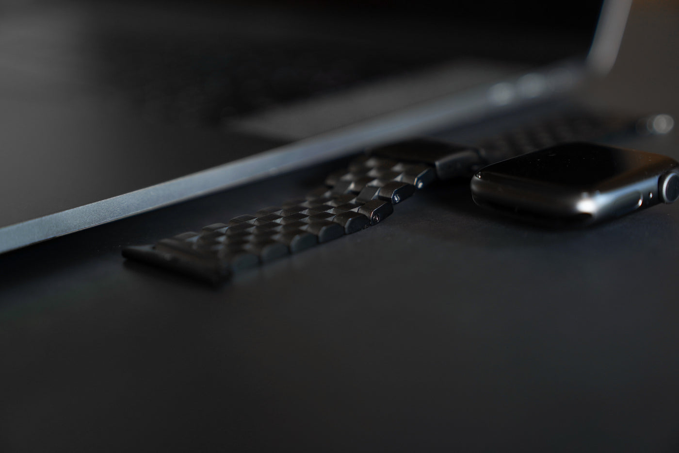 Black smartwatch with a faceted metal link band lying beside a laptop keyboard on a dark desk.