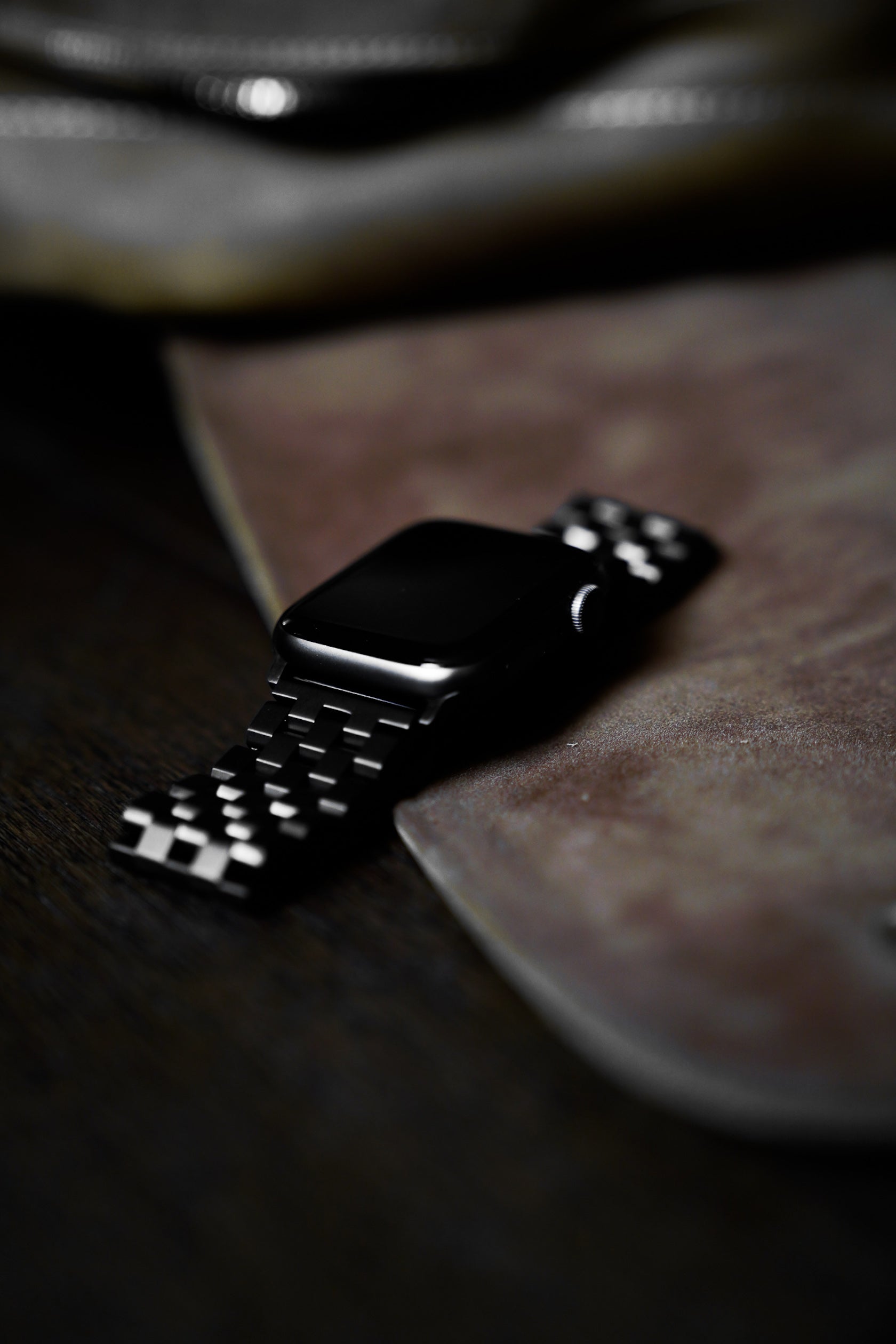 Black metal smartwatch with link-style band resting on a wooden surface beside a leather sleeve