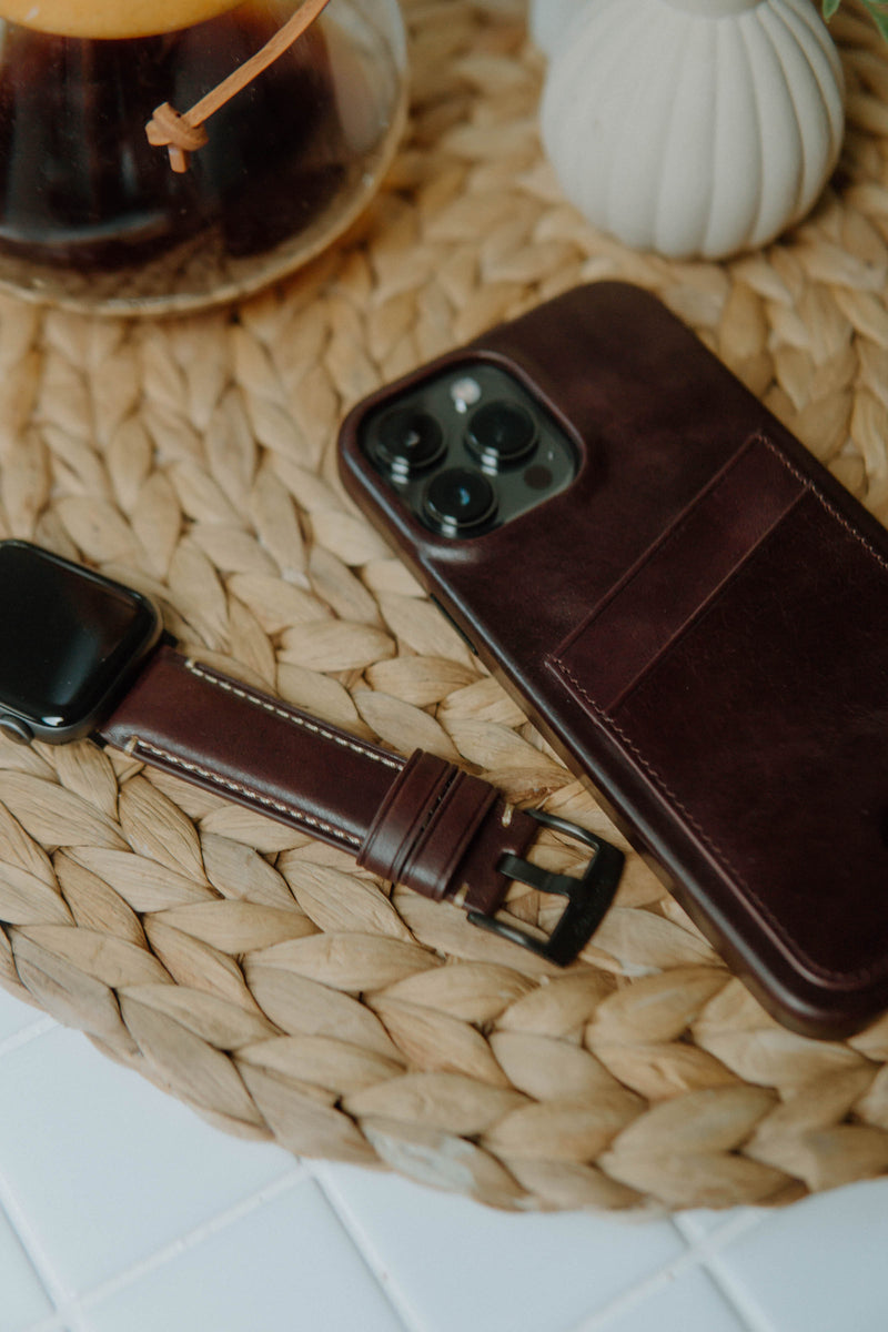 A leather Apple Watch strap next to a phone case on a woven surface.