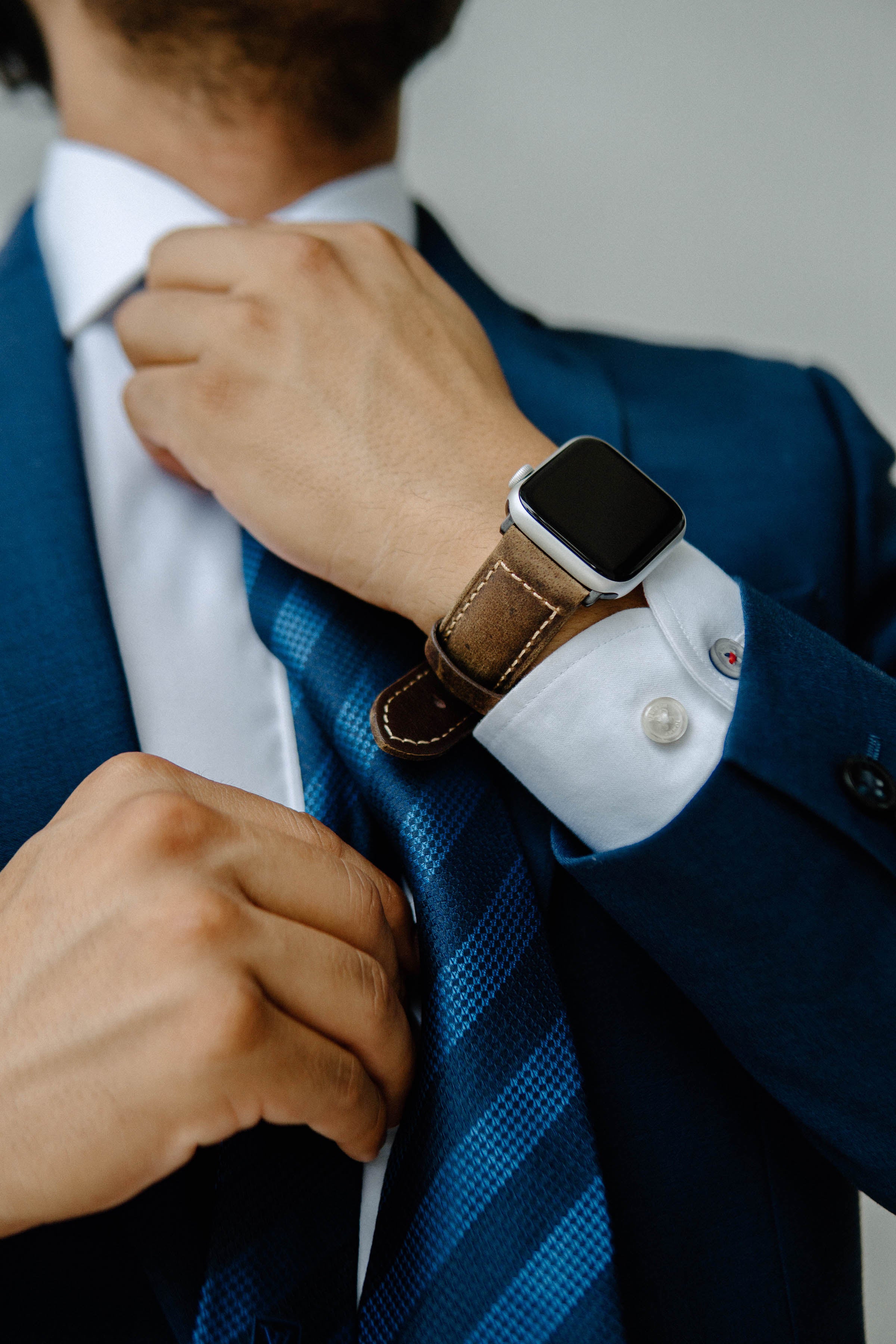 Person in a navy suit adjusting a blue patterned tie, wearing a smartwatch with a brown leather band.
