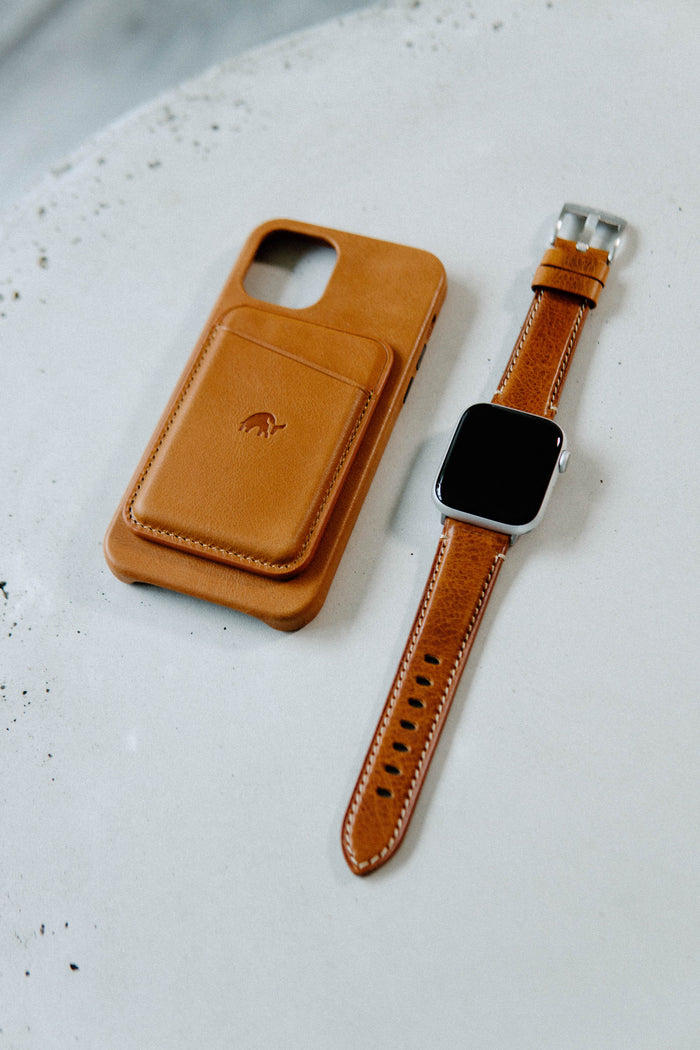 Tan leather smartphone case with stitched card pocket beside matching tan leather smartwatch band and silver square watch face.