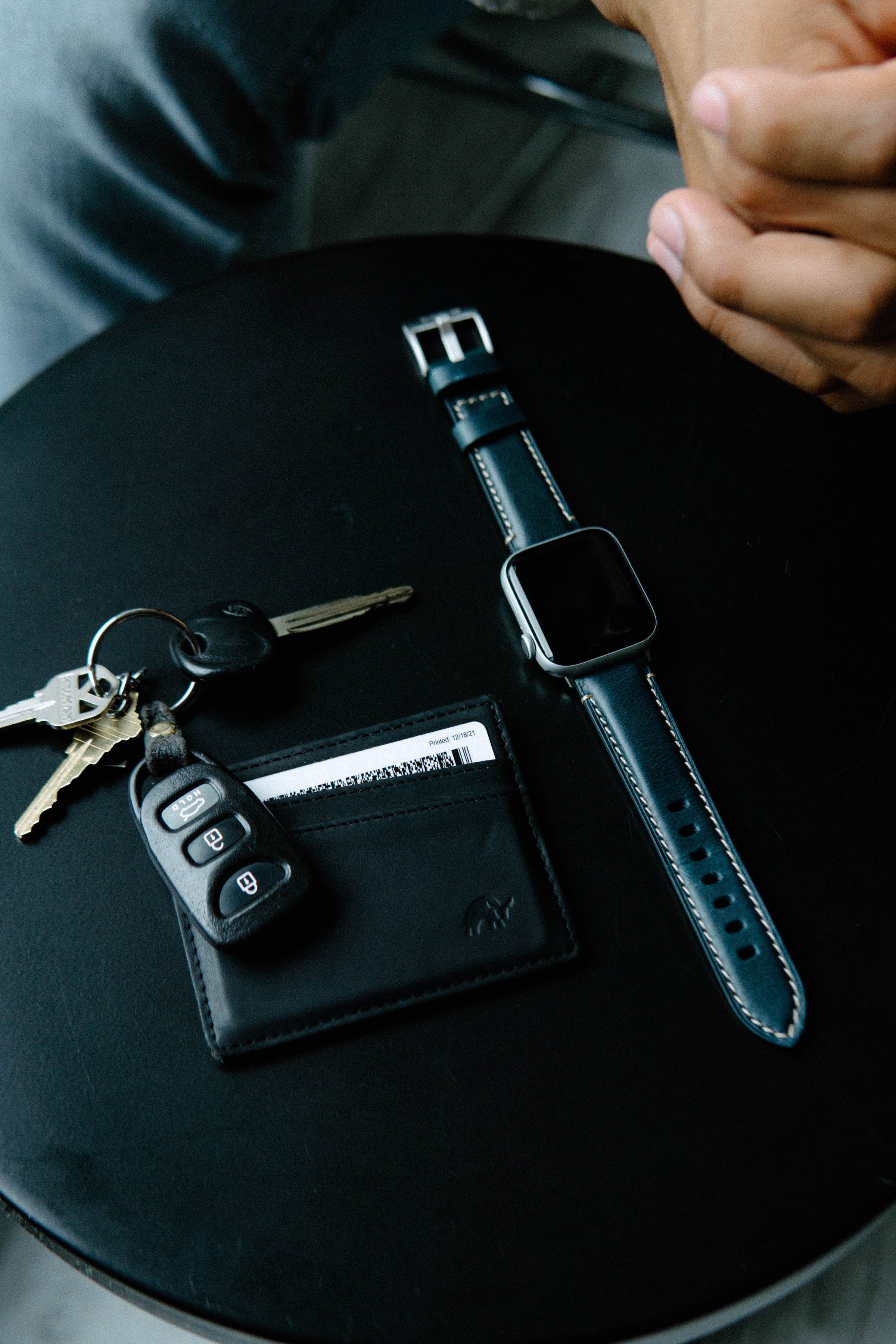 Black card holder showing 'Printed: 12/18/21', car keys with fob, and blue-leather smartwatch on round black table