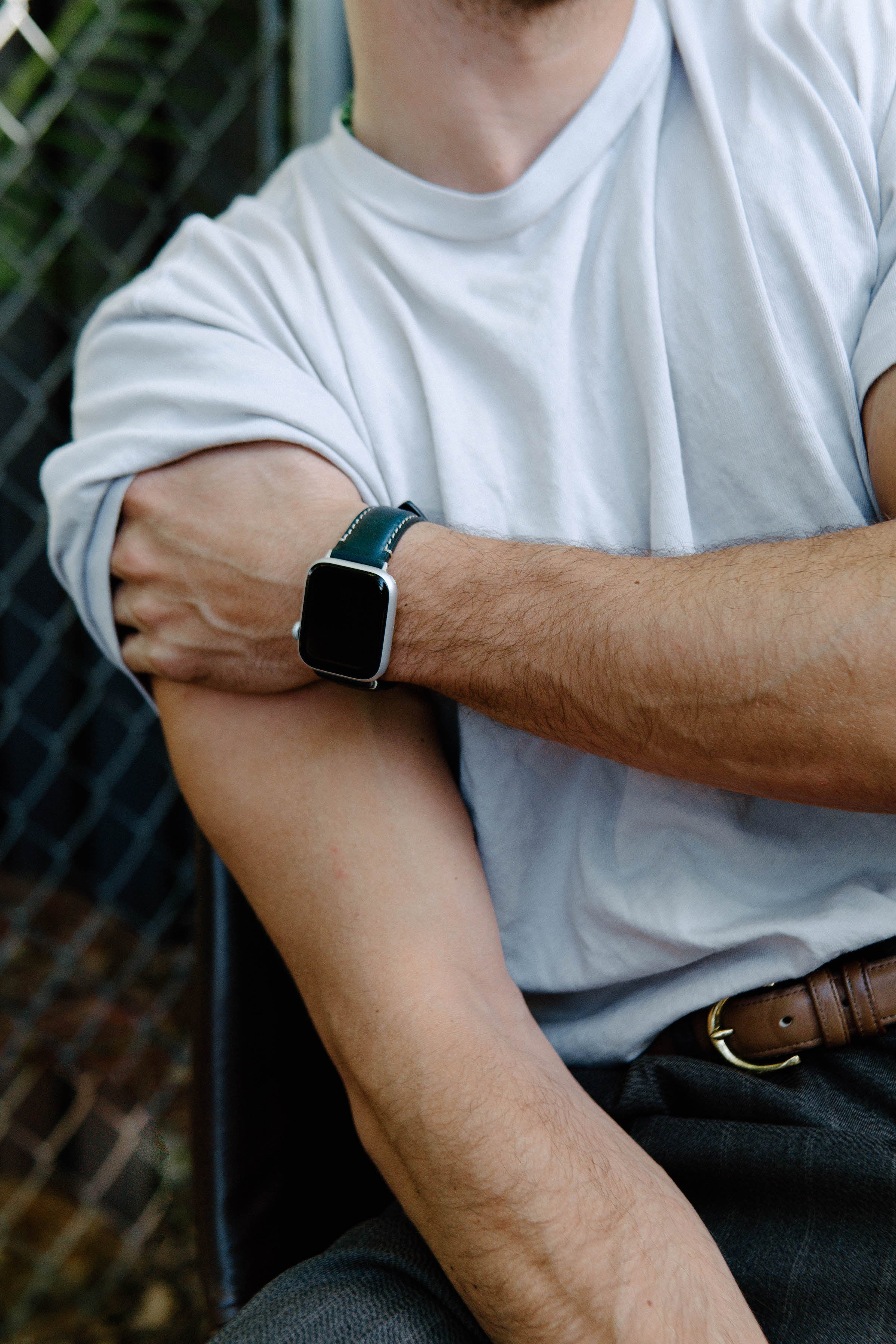 Person wearing a white T-shirt with rolled sleeve and leather-banded smartwatch with dark screen on left wrist, arms crossed