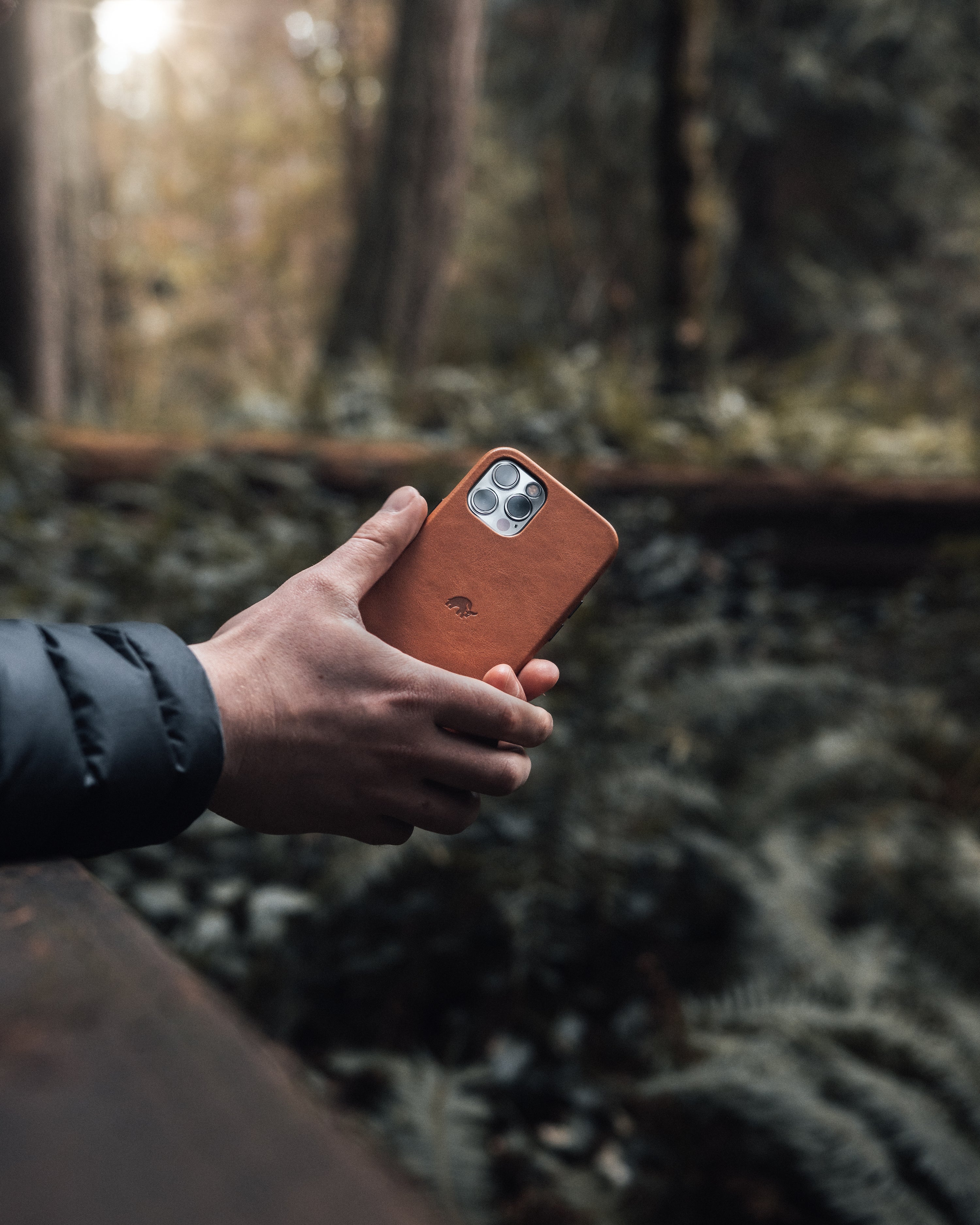A hand holding a leather phone case in a forest setting with ferns in the background.