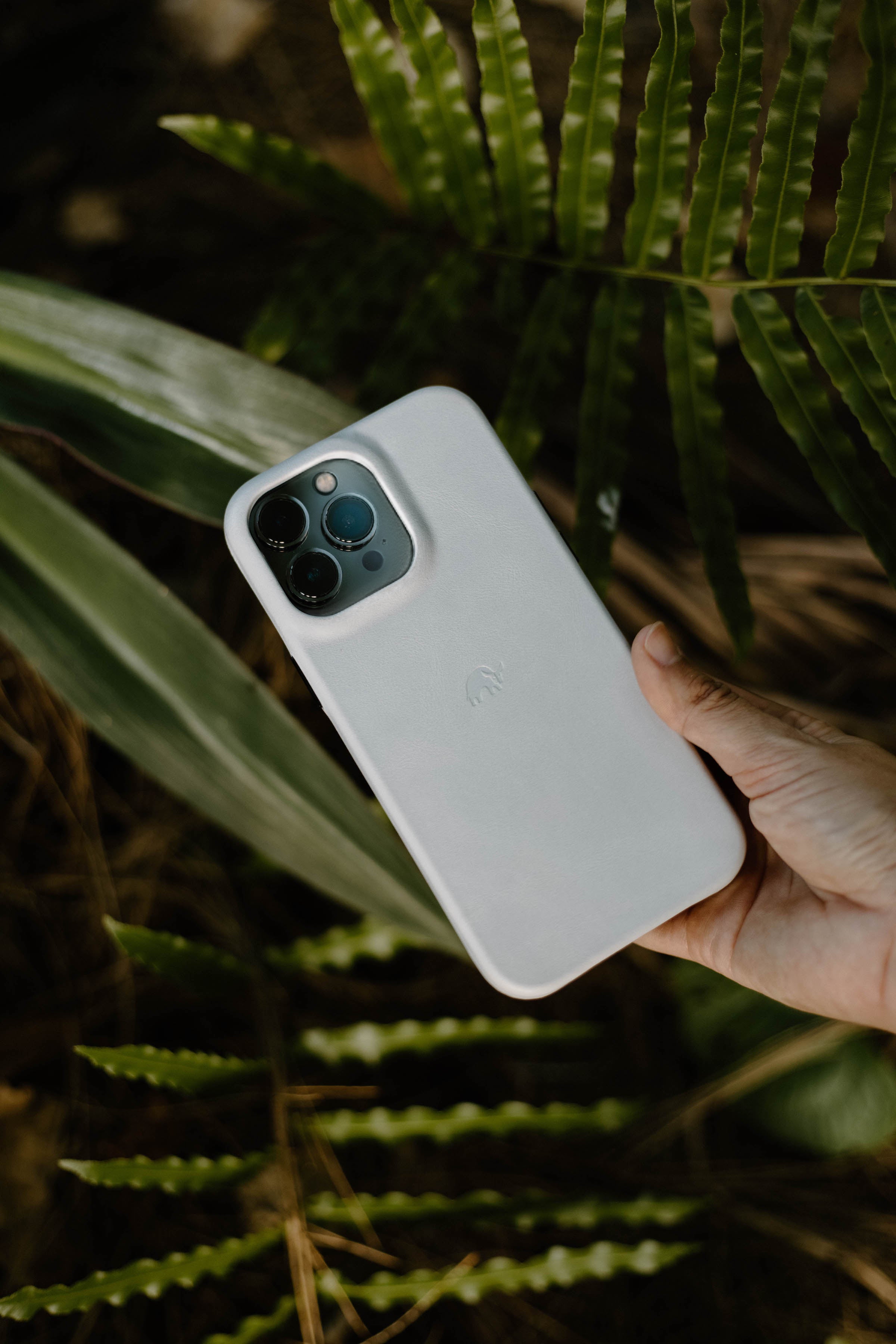 A hand holding a white iPhone case surrounded by green leaves.