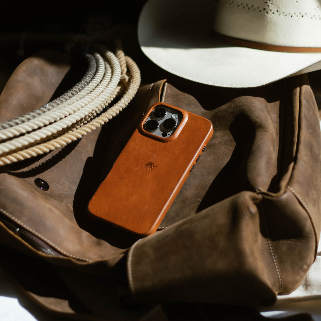 Brown leather phone case on a worn leather jacket beside a coiled rope and white cowboy hat