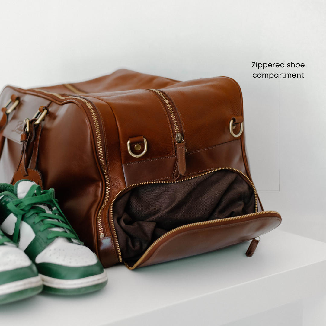 Brown Bullstrap Odyssey travel bag with front Zippered shoe compartment open and green sneakers beside.