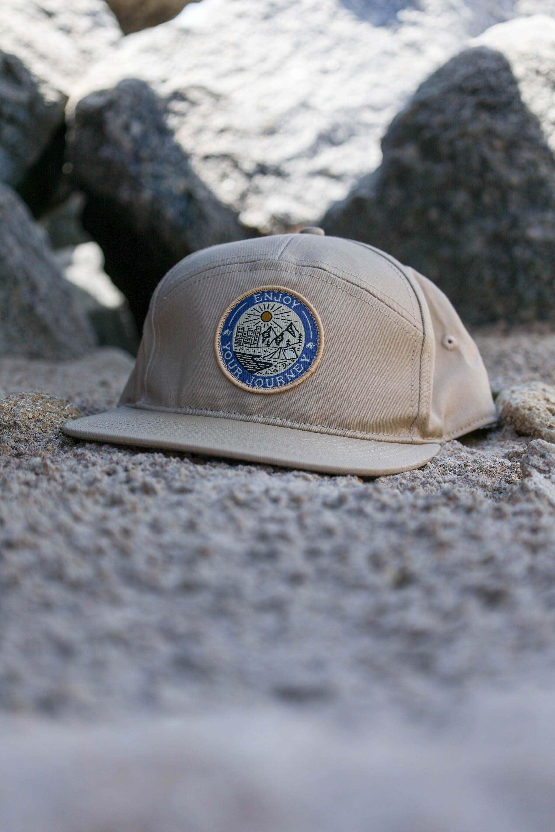 Tan cap with 'ENJOY YOUR JOURNEY' badge on a rocky background.