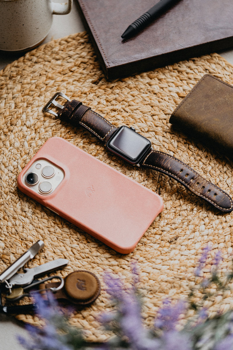 Flat-lay of everyday carry: pink-cased smartphone, leather-strap smartwatch, brown wallet and keys on woven mat with lavender