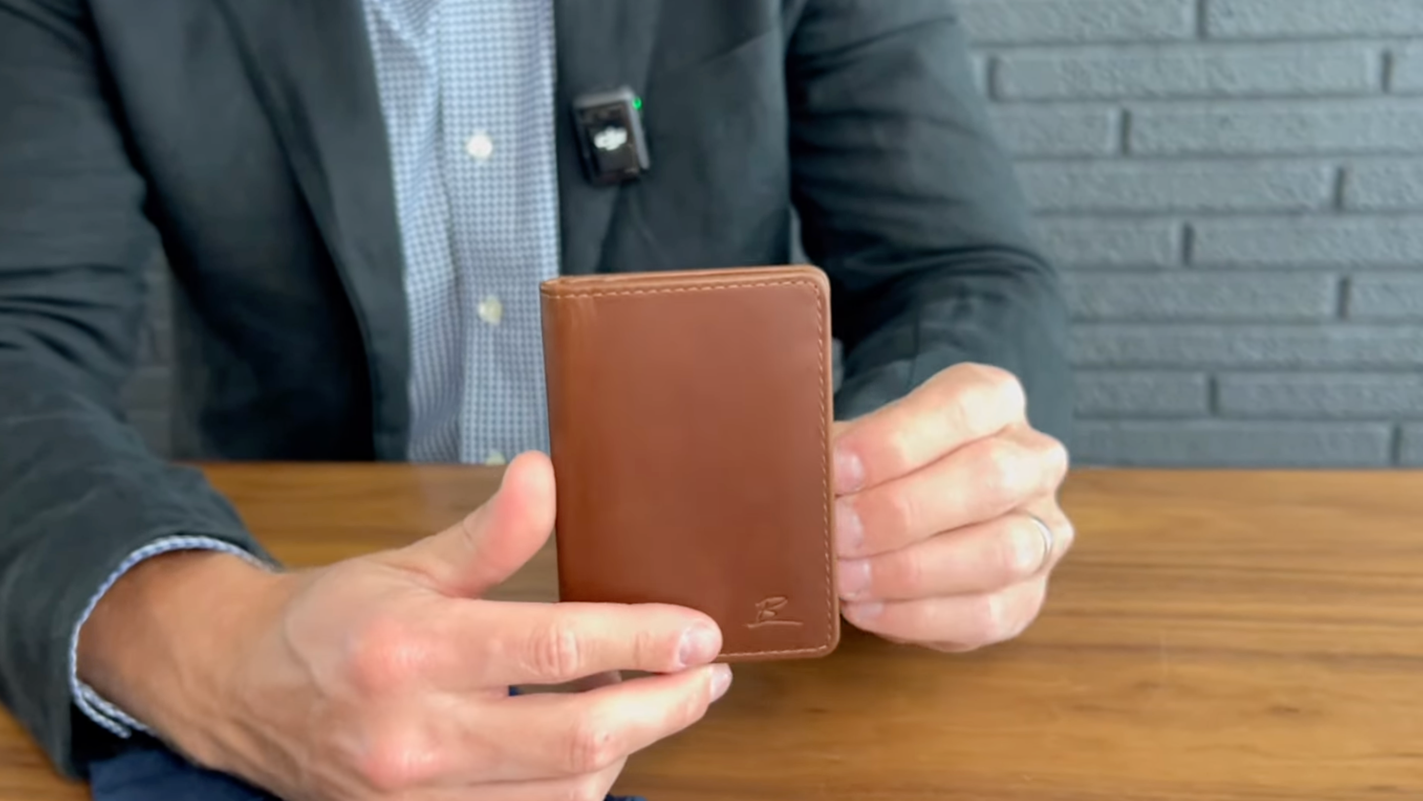 Hands holding a vertical brown leather card wallet above a wooden table, person wearing a blazer visible behind.