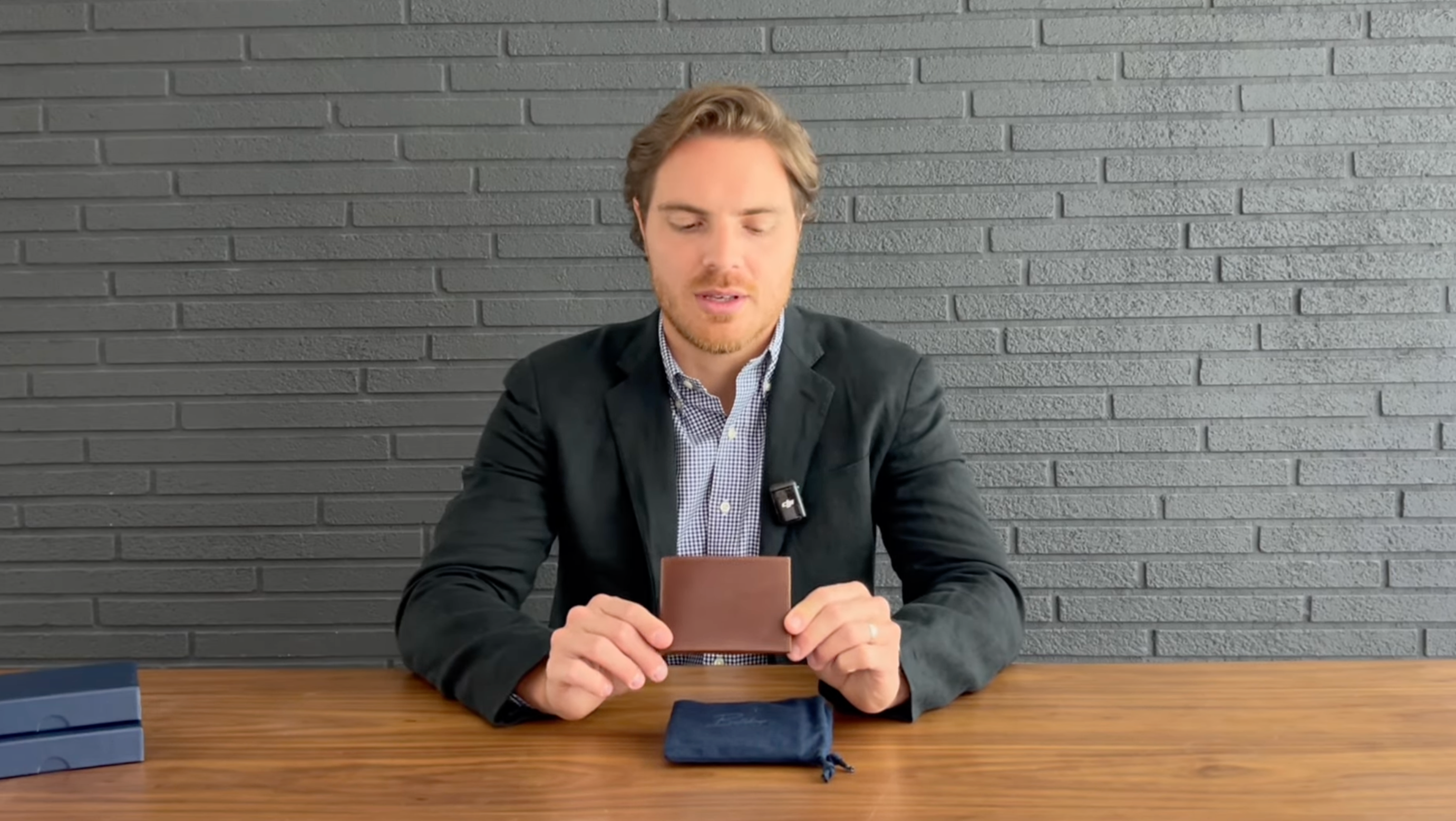 Person wearing a blazer holding a brown leather wallet above a navy pouch on a wooden table, gray brick wall background.