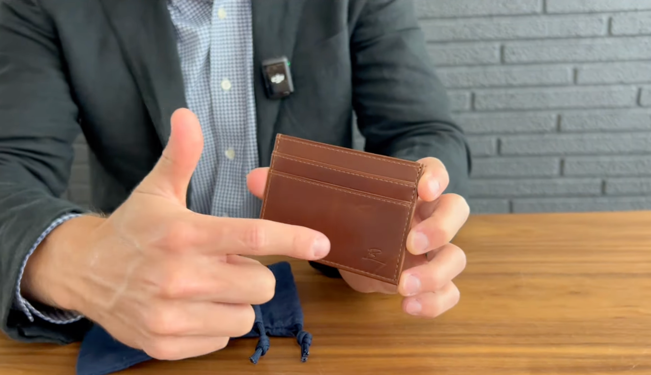 Hands holding a brown leather card wallet, index finger pointing to the front card slot; navy pouch on wooden table below.
