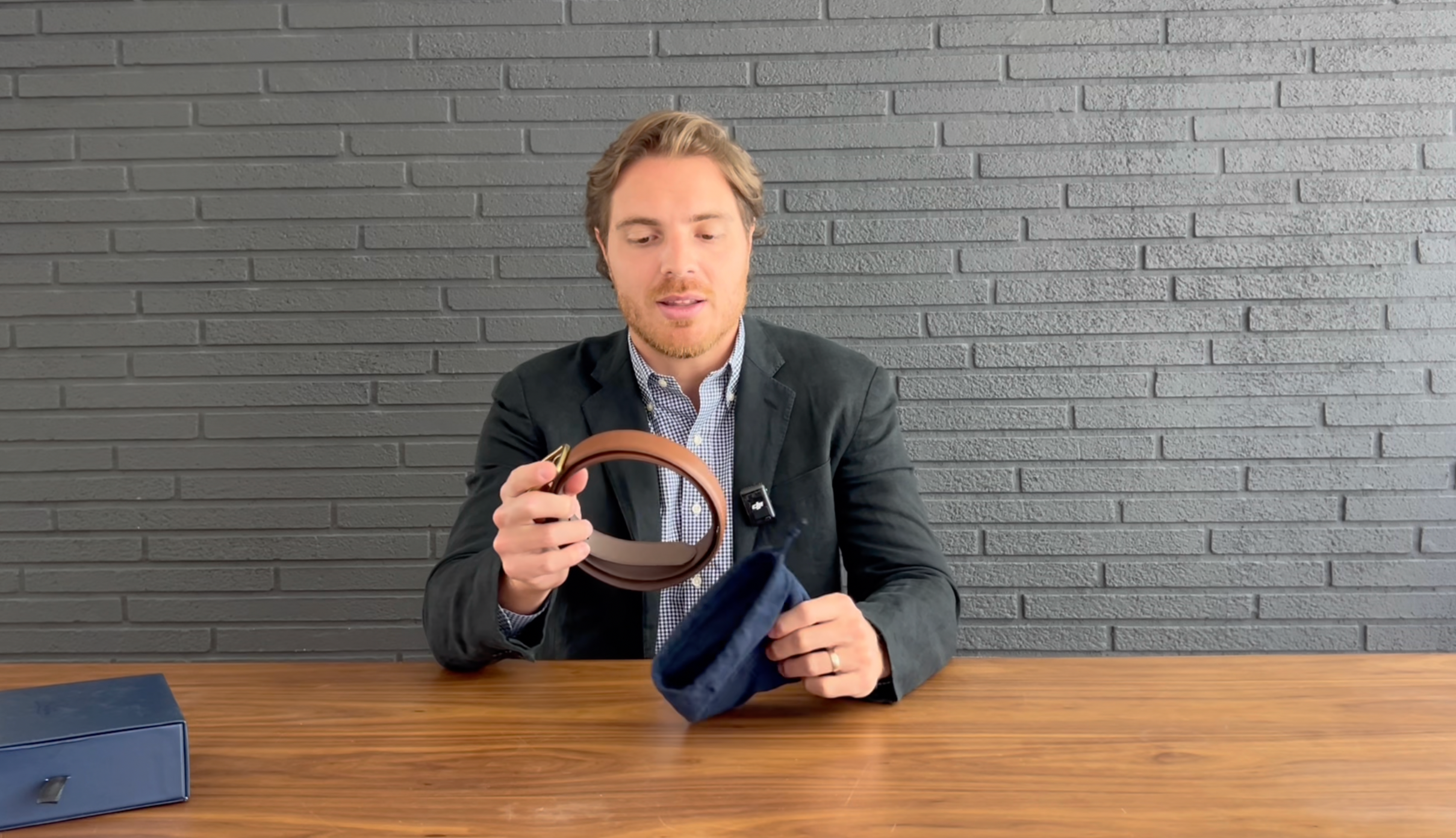 Man at table holding a brown leather belt and a blue suede pouch, gray brick wall background