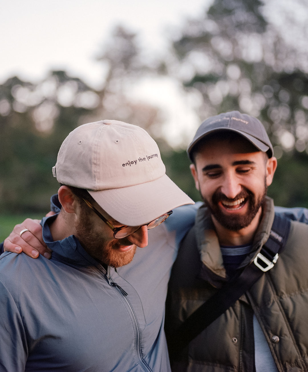 Two men smiling, one wearing a beige hat with 'enjoy the journey' text, the other in a dark hat.