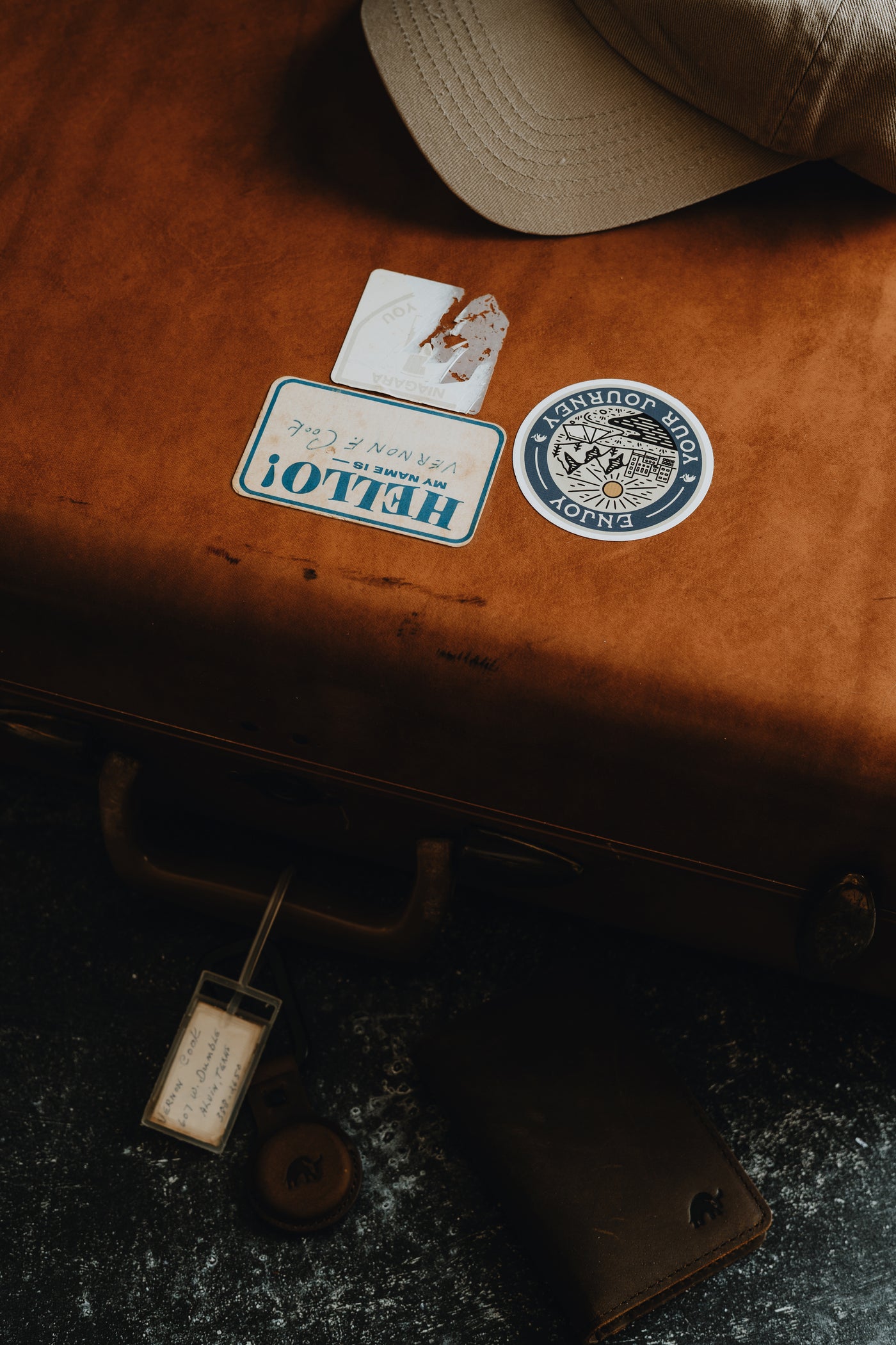 A brown suitcase with various stickers, a hat, and a wallet on a dark surface.