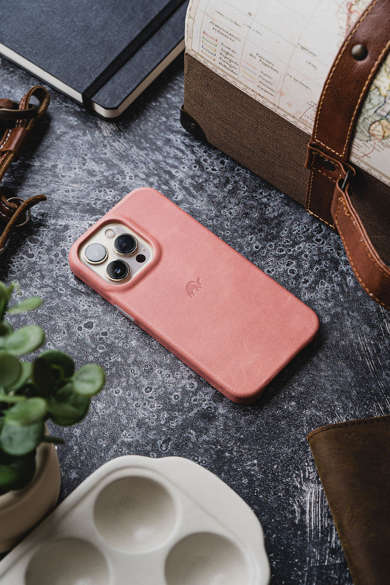Pink leather iPhone case with triple camera on a textured dark tabletop, beside a notebook, leather travel bag, and potted plant.