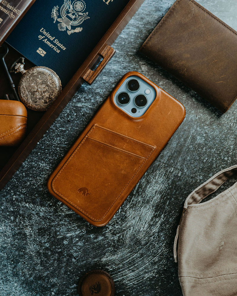 Leather brown wallet-style phone case on tabletop with passport reading 'United States of America', pocket watch and canvas bag.