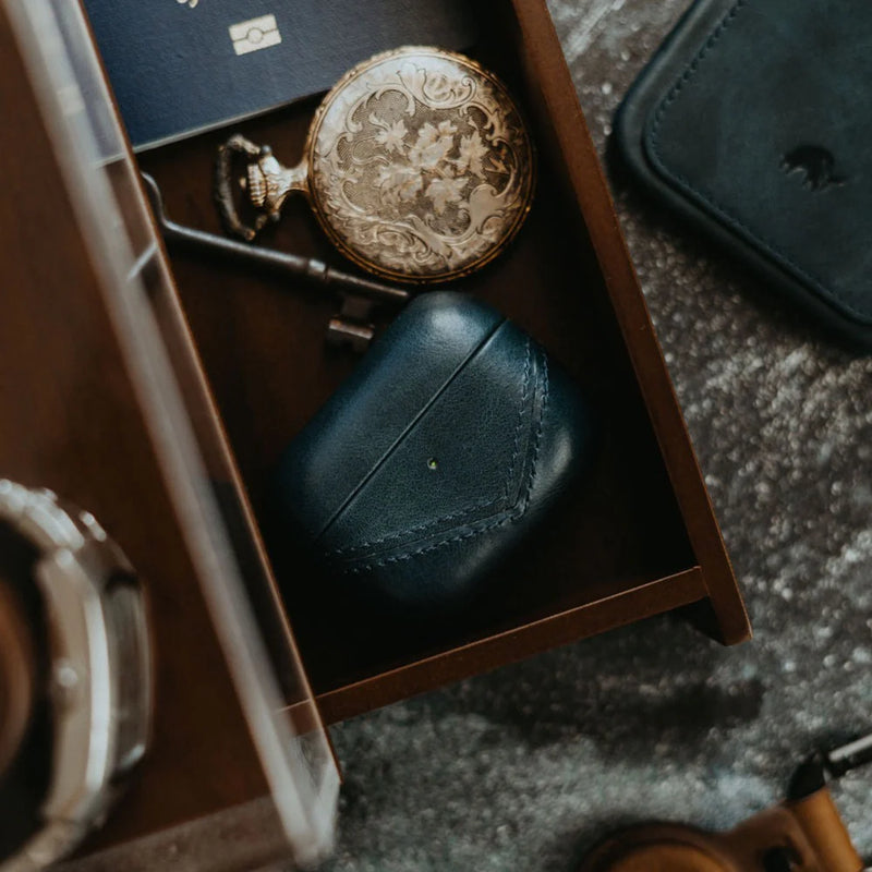 Top-down view of Terra leather AirPods case with V-stitch detail and front LED hole in a wooden drawer with pocket watch