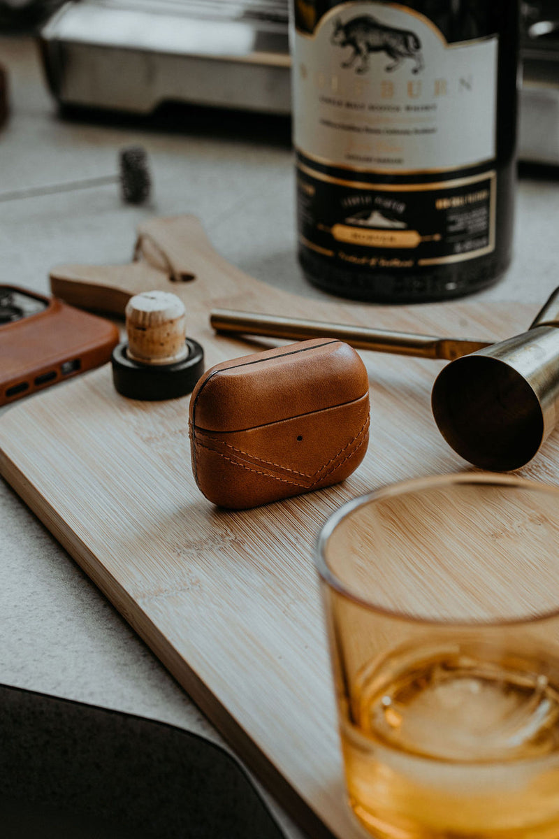 Leather AirPods case on a wooden surface with a drink and accessories