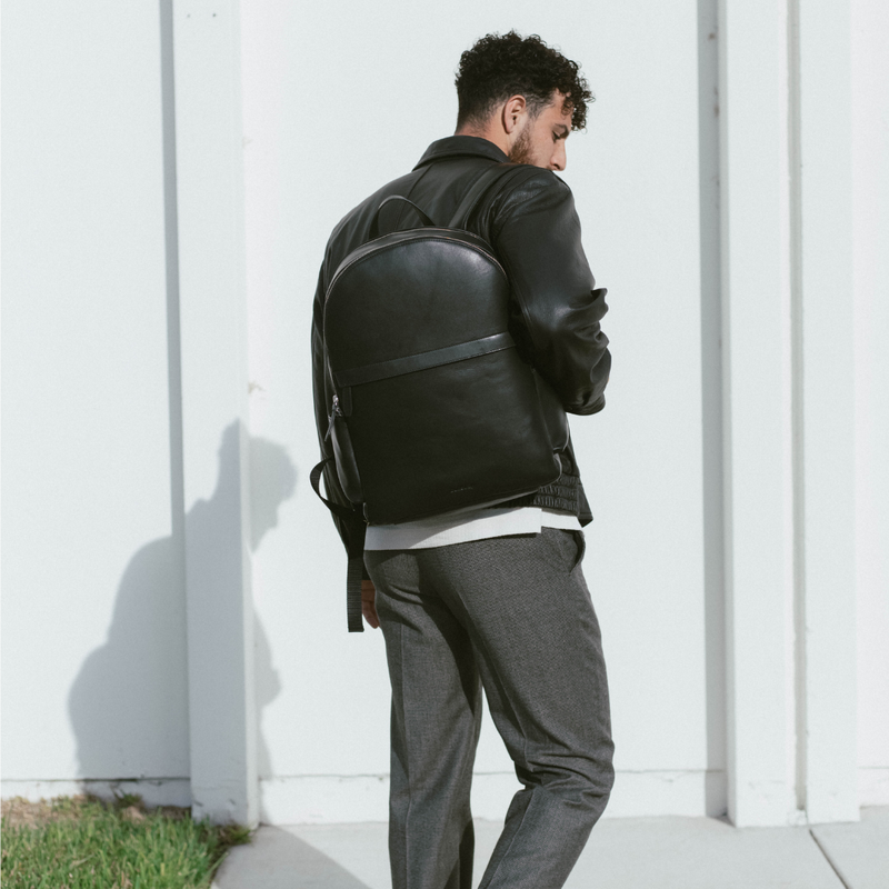 Man in black leather jacket with a black leather backpack, back turned and looking over his shoulder by a white wall.