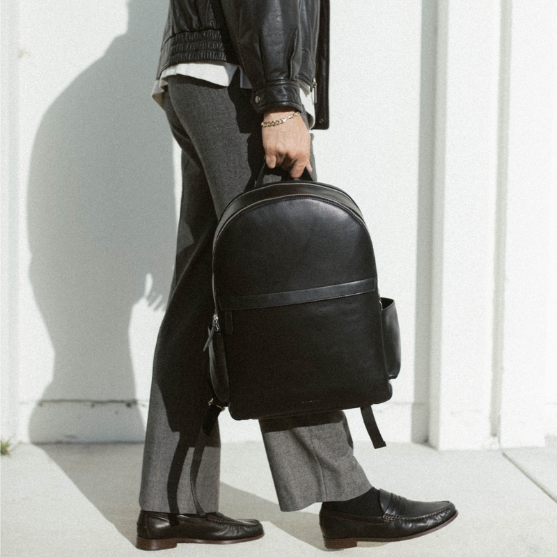Person walking, holding a black leather backpack by its top handle; wearing gray trousers, black loafers and leather jacket.