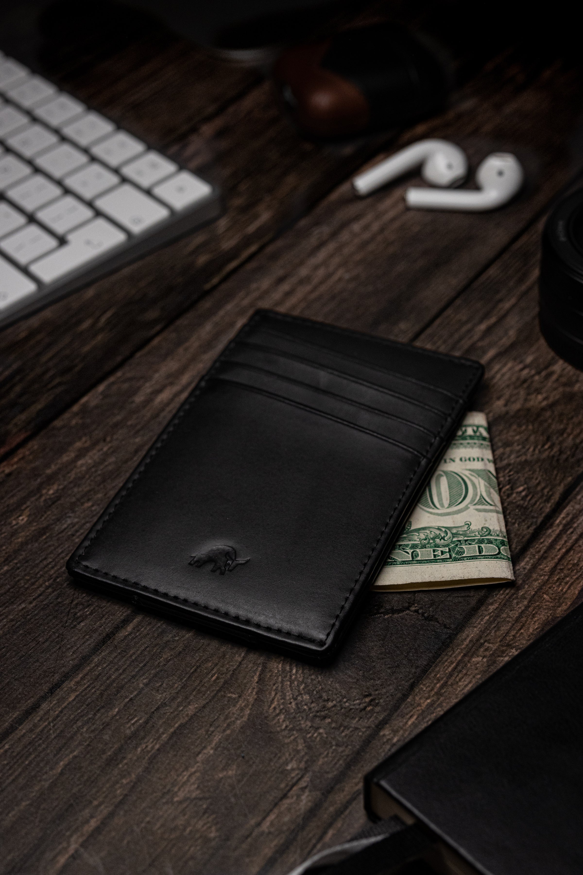 Black leather card wallet on wooden desk with a folded one-dollar bill showing 'ONE' and 'IN GOD', keyboard and earbuds.