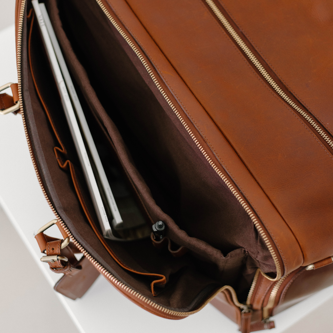 Open brown leather travel bag interior with padded laptop sleeve, pen holder, and zippered pocket.