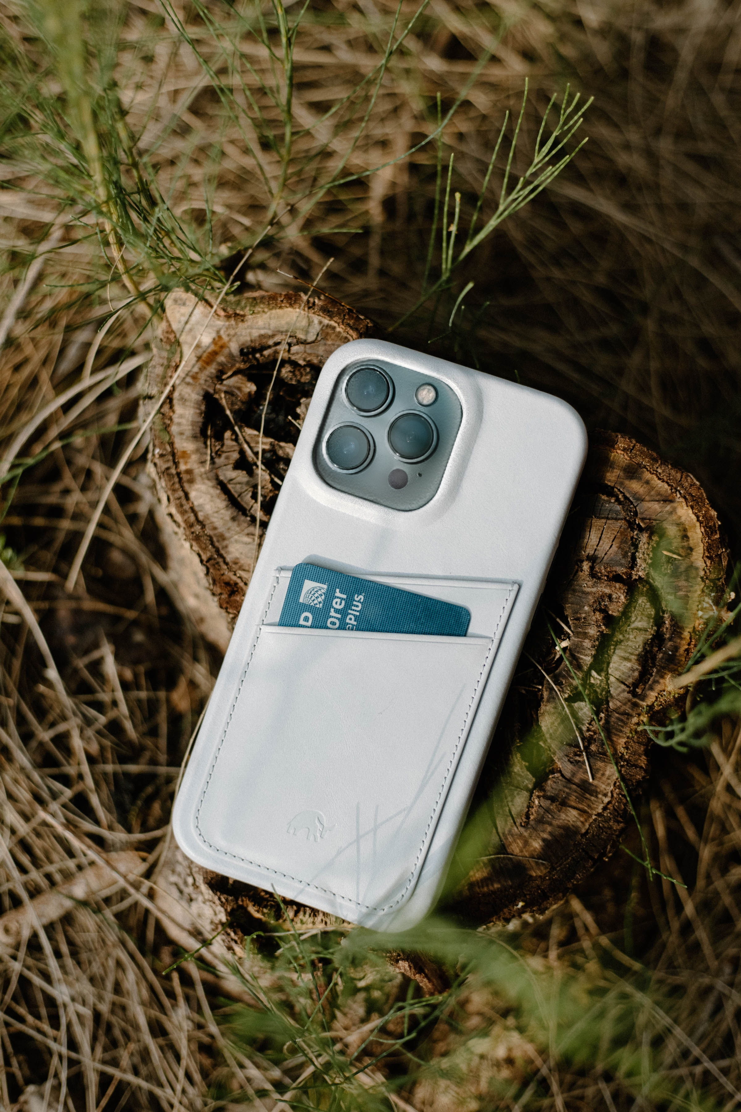 White phone case with a blue card labeled "D rover plus" tucked in the back pocket, resting on a tree stump