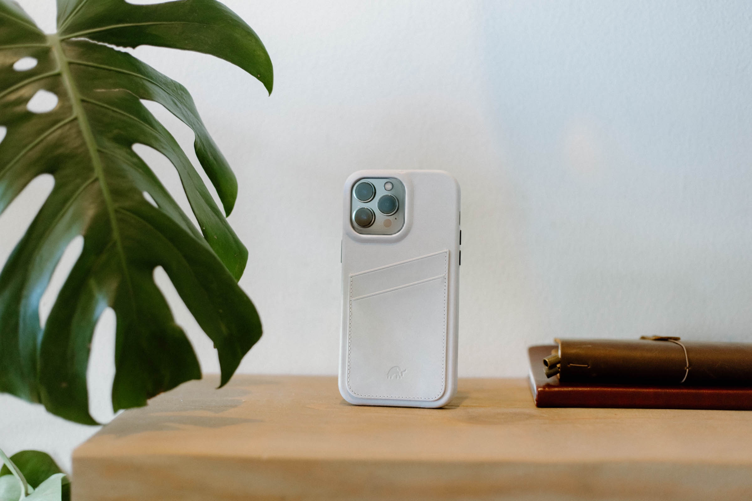 White phone case with card pocket on a wooden shelf flanked by a large green monstera leaf and leather notebooks.