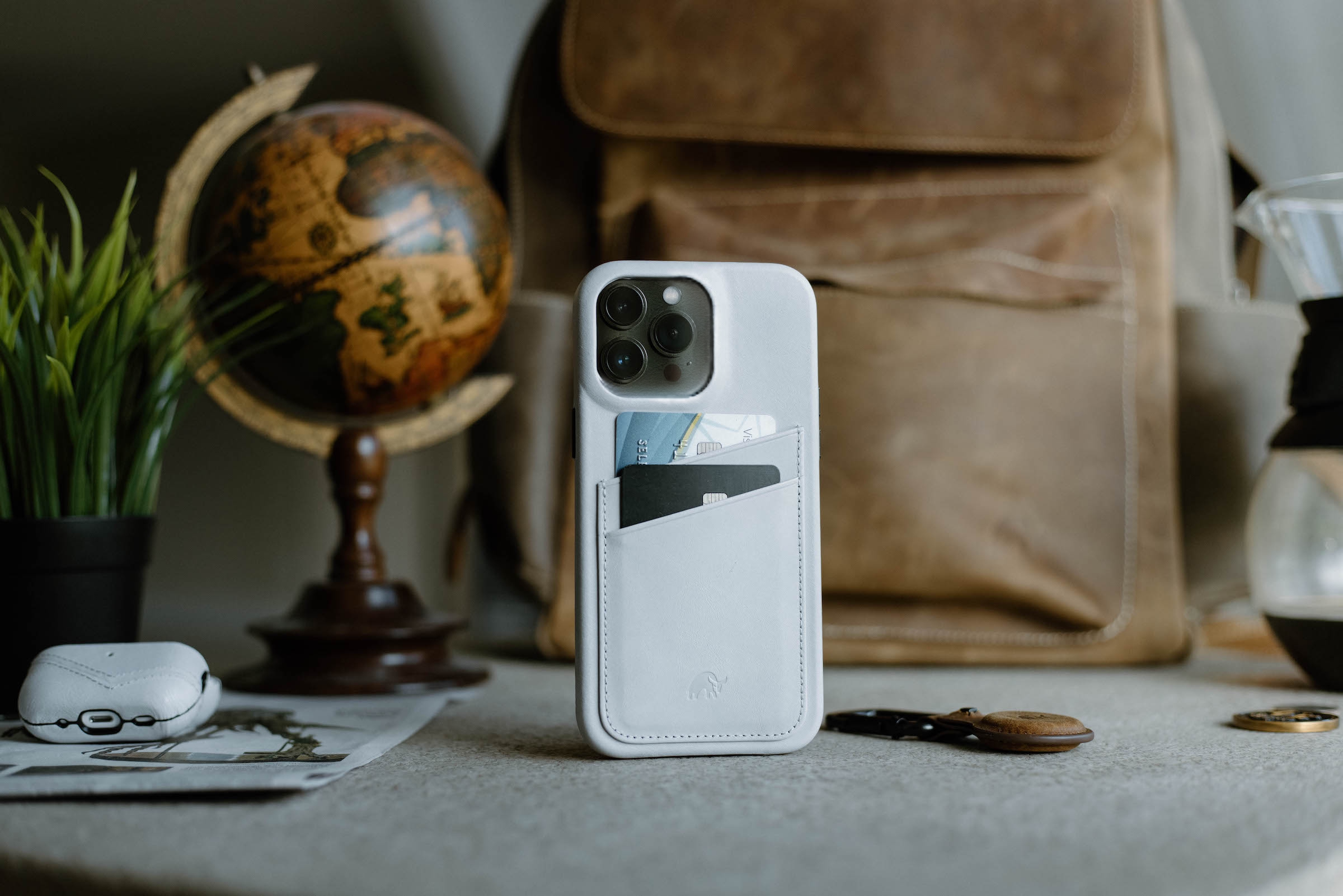 White leather iPhone case with two cards in back pocket, on table with globe and leather backpack blurred behind.