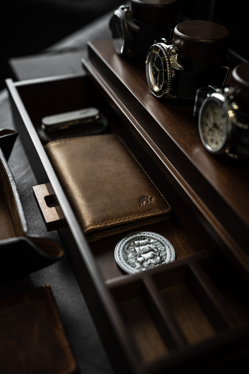 Angled overhead view of closed brown leather Messenger Wallet showing stitched edge and embossed logo inside a wooden tray with watches and a coin