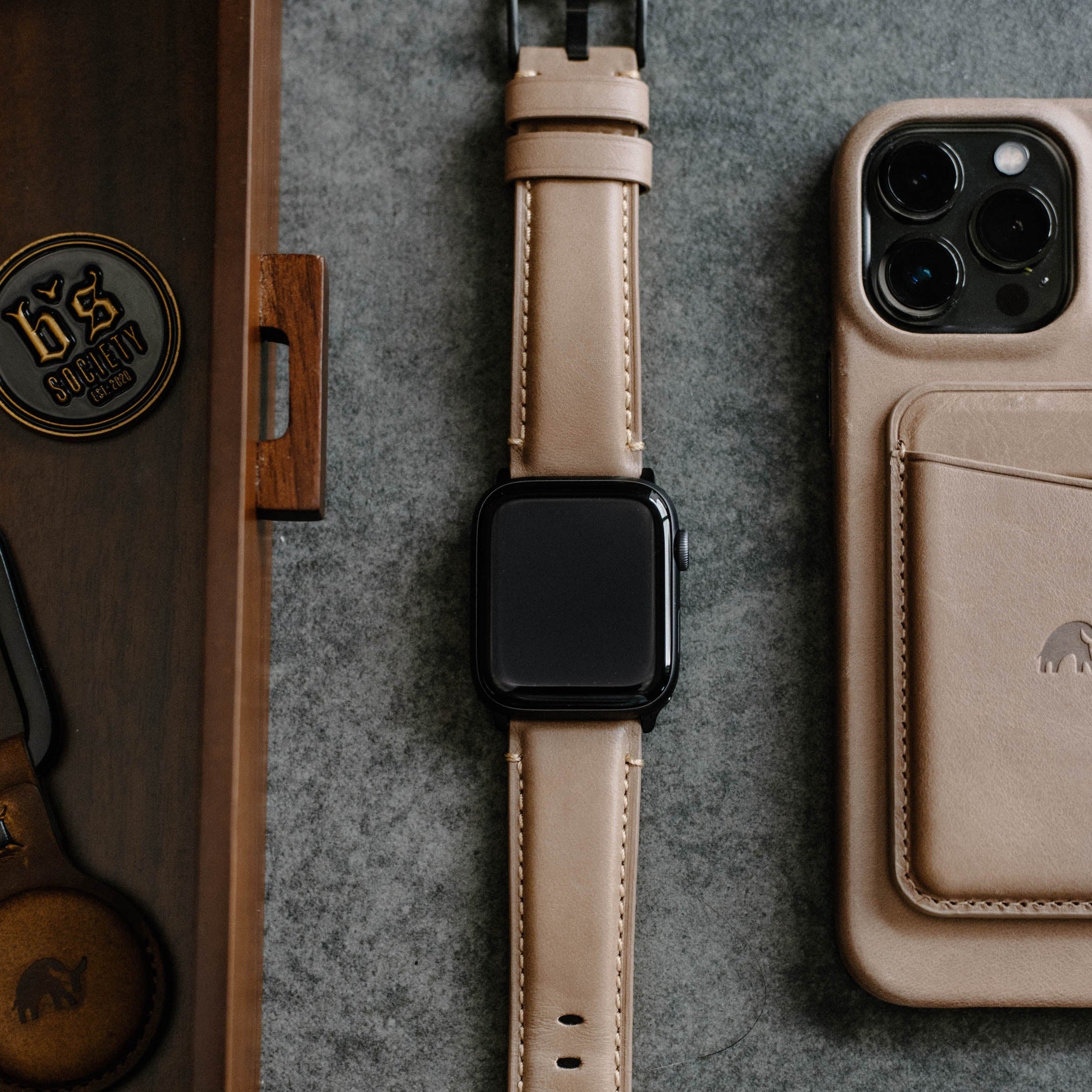 Leather Apple Watch Strap in Dune color displayed on a gray surface next to a phone.