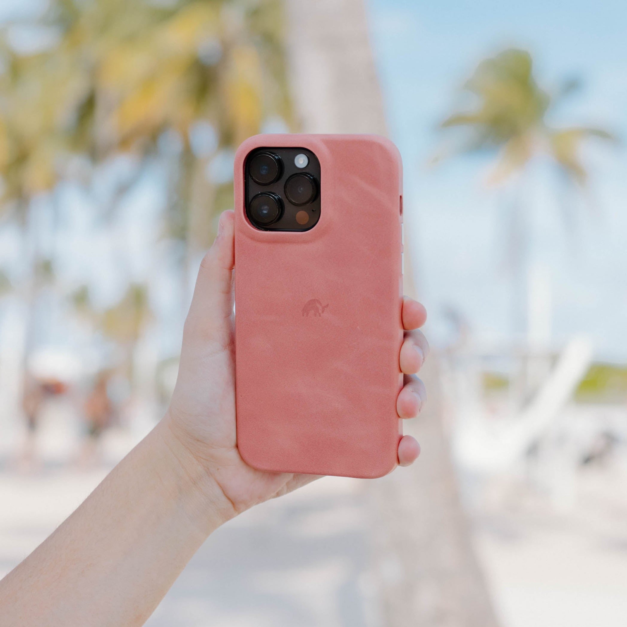 A person holding a pink Minimalist Case for iPhone 16 in a beach setting with palm trees in the background.
