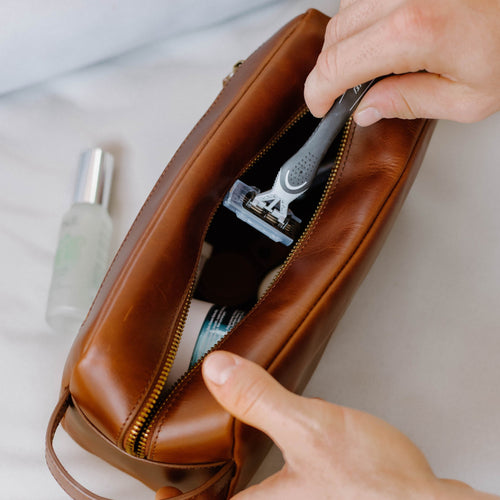 Hand placing a razor into a brown leather toiletry (dopp) bag with a visible skincare bottle and tube.